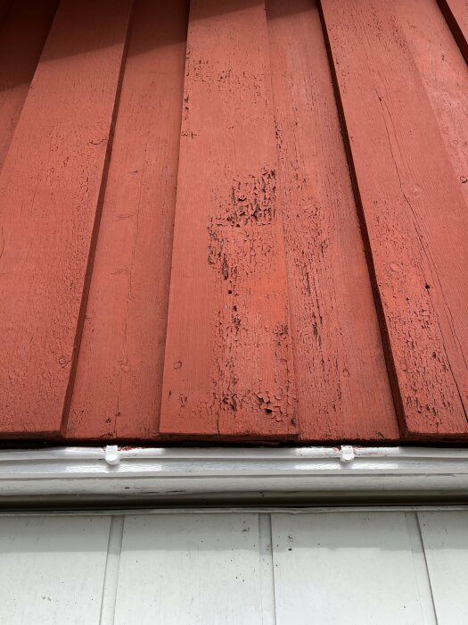 Närbild på en röd träfasad med flagnande färg och sprickor, över en vitmålad hängränna.