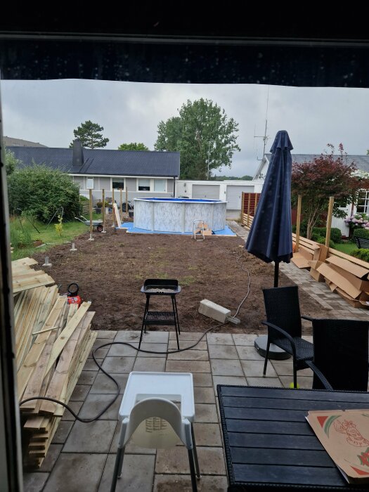 Bakgård under renovering med oavslutad pool, trästaplar, utemöbler och en grill på en stenlagd yta. Hus och trädgård i bakgrunden.