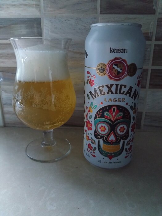 En burk med Keisari Mexican Lager och ett glas öl med skumkrona placerade på ett köksbord framför en kakelvägg i ljusa nyanser.