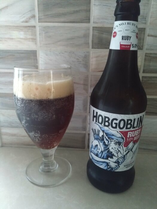 En flaska Hobgoblin Ruby Beer bredvid ett glas fyllt med rubinfärgad öl med skum, på en köksbänk mot en kaklad vägg.