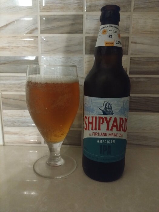 Flaska Shipyard American IPA och ett glas med samma öl på köksbänken. Bakgrund i kakel med jordnära nyanser.