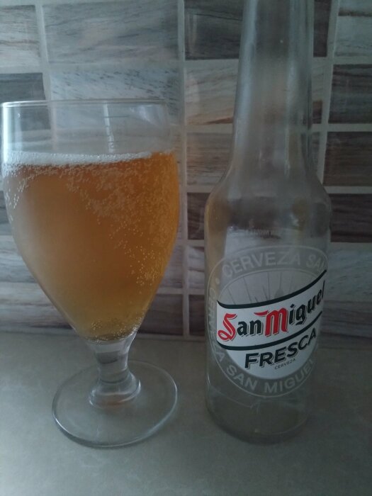 Ett glas fyllt med San Miguel Fresca öl och en tom San Miguel Fresca ölflaska står på ett bord med en kaklad bakgrund.