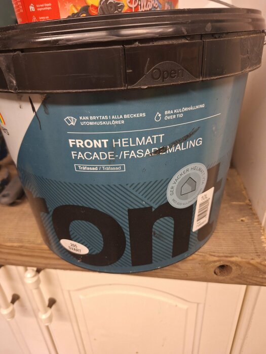 En burk fasadfärg från märket Front Helmat med etiketten "Facade-/Fasadfärg" stående på en trähylla. Färgen är svart, 10 liter.