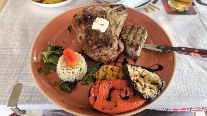 En T-bone steak på en tallrik med en klick smör på toppen, serverad med grönsaker, grillad paprika, aubergine och en risboll, tillsammans med ett glas öl i bakgrunden.