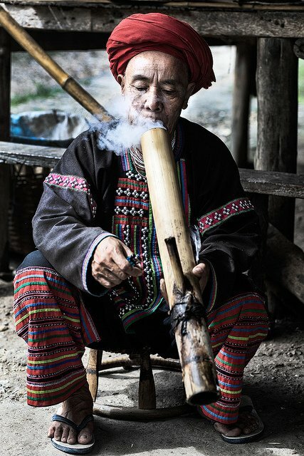 Person klädd i färgglada traditionella kläder röker en bambupipa och sitter på en pall.