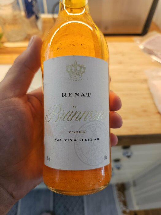 En hand håller i en flaska "Renat Brännvin" vodka från V&S Vin & Sprit AB. Flaskans etikett är vit med guldfärgad text och en krona.