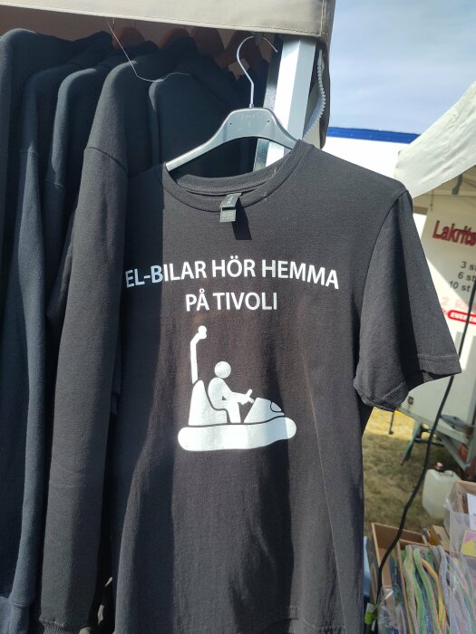 Svart t-shirt med texten "EL-BILAR HÖR HEMMA PÅ TIVOLI" och en illustration av en person som kör en radiobil.