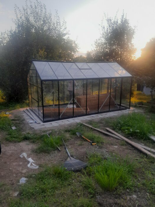 Nybyggt växthus av glas i en trädgård, med solnedgång i bakgrunden och träd samt trädgårdsredskap runt om.