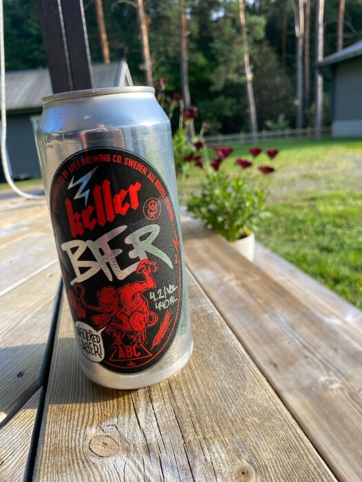 En burk Keller bier från Apex Brewing Co. på ett trädäck i solsken, med en trädgård och skog i bakgrunden.