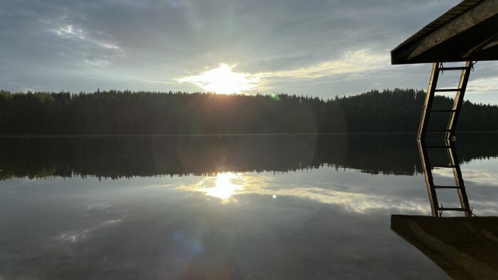 Solen går ner över en skogssjö med stilla vatten som speglar himlen och träden, medan en brygga och stege syns till höger i bilden.