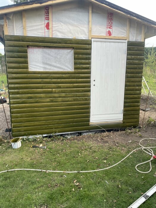 Ett halvfärdigt trähus med gröntjära på ytterpanelen, ett vitt fönster som är täckt och en vit dörr. Verktyg och en burk med färg ligger på marken framför huset.