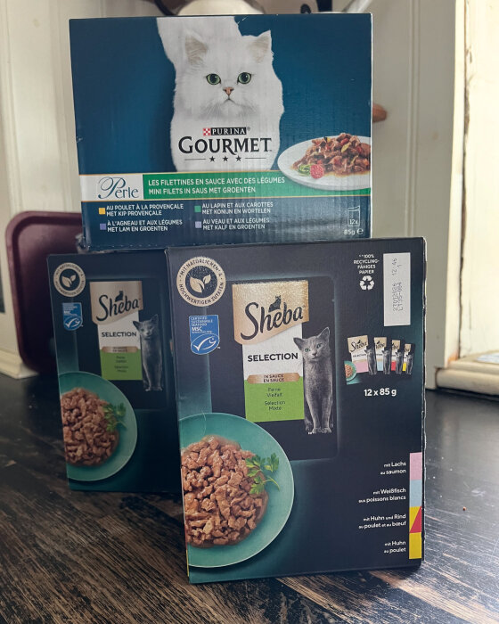 Förpackningar med kattmat av märkena Purina Gourmet och Sheba placerade på ett köksbord.