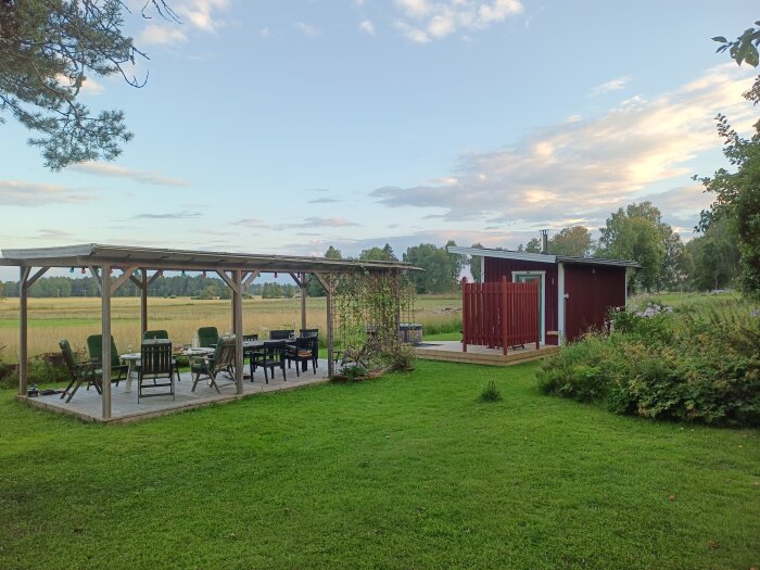 Plank byggt för en utedusch utan överliggare. Bredvid står en altan med tak och utemöbler i en trädgård. Landskap med äng i bakgrunden.