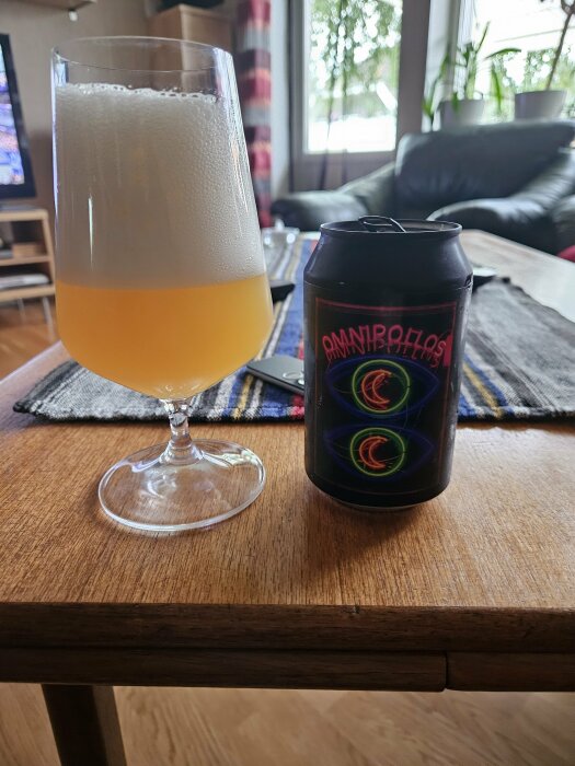 Ett glas fyllt med öl från Omnipollo med rik skumkrona står på ett bord bredvid en svart Omnipollo-burk.