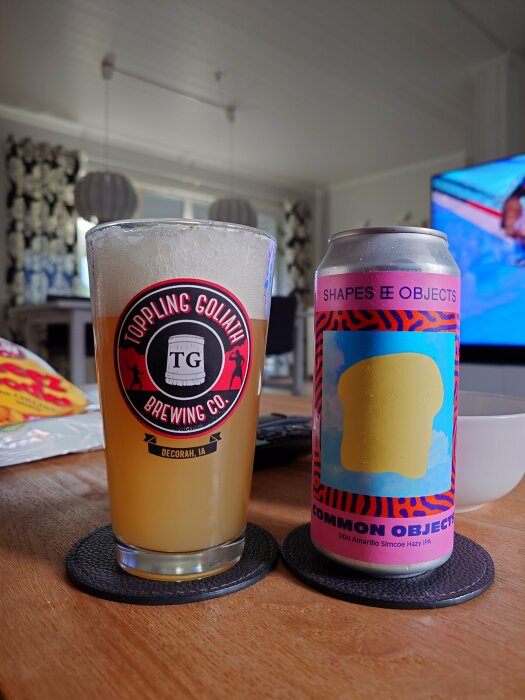 Ett glas öl från Toppling Goliath Brewing Co. bredvid en färgglad burk Shapes & Objects Common Objects DDH Amarillo Simcoe Hazy IPA, stående på ett bord.