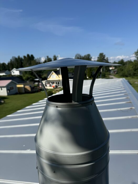 Skorstenshatt på plåttak i solsken med hus och träd i bakgrunden. Skorstenshatten har en fyrkantig plåt som skydd ovanpå.