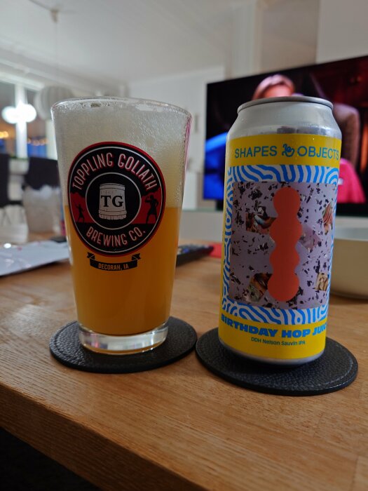 Glas med Toppling Goliath-logga och ölburk från Shapes & Objects med texten "Birthday Hop Juice" DDH Nelson Sauvin IPA står på ett bord inomhus.