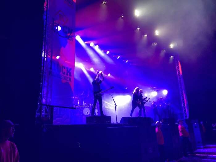 Rockband uppträder på scen med färgglada ljuseffekter och en bannér med texten "Rock Klassiker" i bakgrunden. Scenvakter syns nedanför scenen.