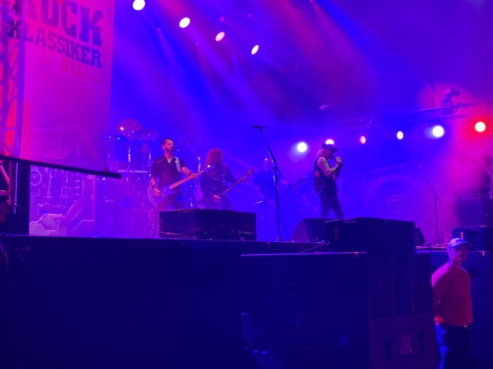 Rockband spelar på scen med lila och blått scenljus. Sångaren står med mikrofonen, gitarrister och trummis i bakgrunden. Säkerhetsvakt står vid scenkanten.