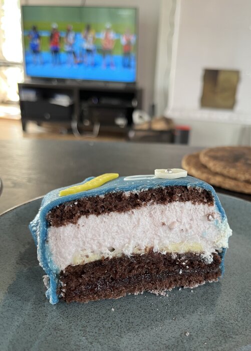 Närbild på en bit tårta med blå marsipan och chokladlager, samt ett TV-apparat som visar ett sportprogram i bakgrunden.