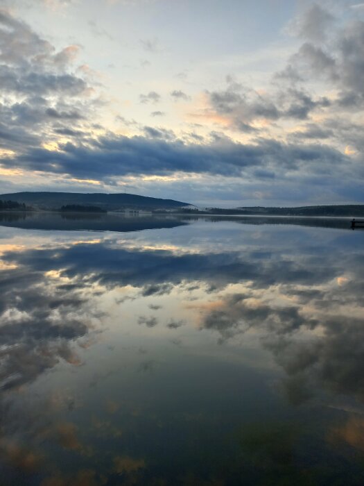 Vy över en stilla sjö med spegelblank yta som reflekterar den molniga himlen vid skymning. Skogsklädda kullar syns i fjärran.