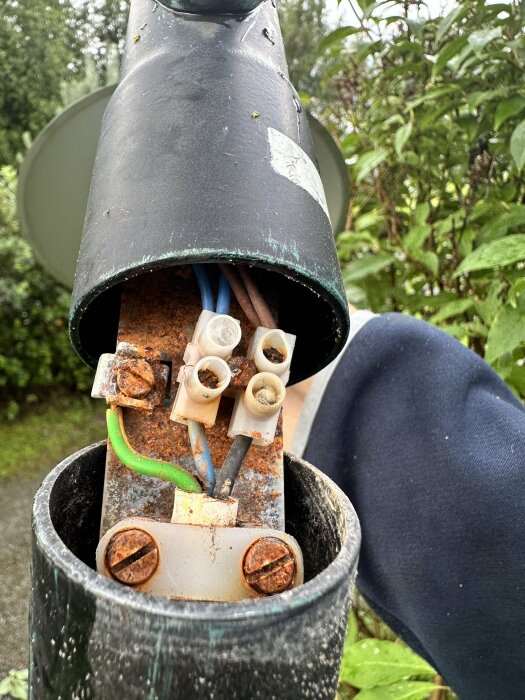 Närbild på en trasig utomhuslampa med rostiga skruvar och exponerade kablar. Bakgrunden visar gröna blad och en trädgårdsmiljö.