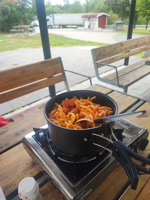 En kastrull med makaroner, wienerkorv och tomatsås från en burk Mutti på en bärbar spis vid ett bord på en rastplats med bänkar och lastbil i bakgrunden.