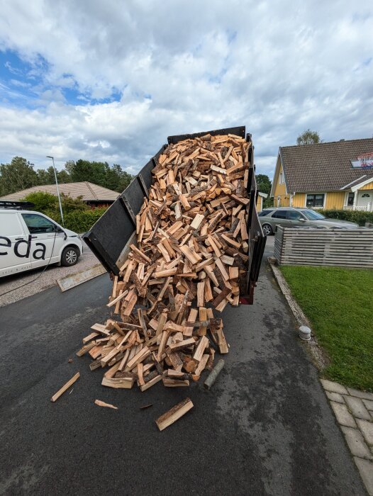 En lastbil tippar av en hög med vedkubbar på en asfaltsväg bredvid ett bostadshus.