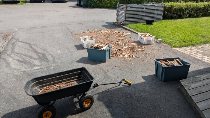 Fyllda plastlådor med ved ligger på gatan bredvid en kärra och en hög med vedflisor och spill, bredvid en plattsatt gång och en gräsmatta.