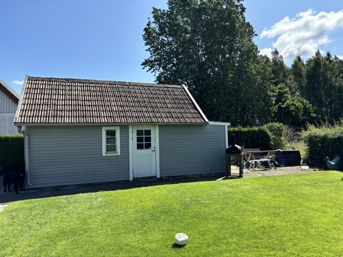 Grått uthus med tegeltak och vit dörr, omgiven av trädgård med gräs och stora träd i bakgrunden, fotboll på gräset.