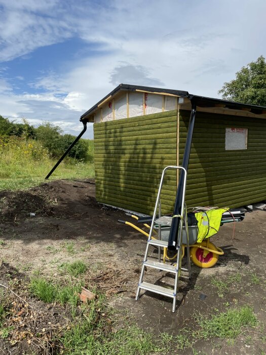 Grönt hus med nyinstallerade svarta stuprör, stege och skottkärra i förgrunden. Klar himmel och grönska i bakgrunden.