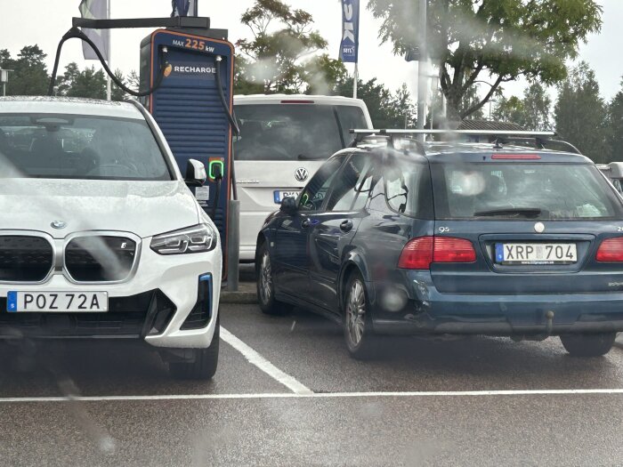 Tre bilar parkerade vid en laddstation. En vit elbil laddas medan en blå bil blockerar en laddningsplats. Träd och skogar syns i bakgrunden.