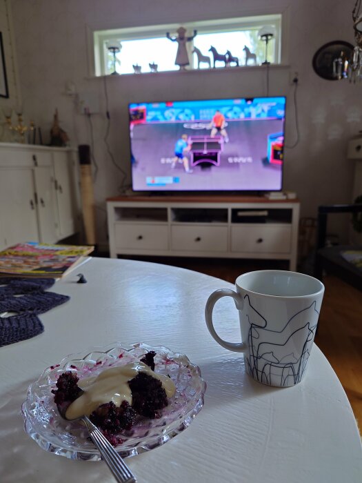 Blåbärspaj med vaniljsås på en tallrik och en kaffekopp på ett vitt bord, med en TV i bakgrunden som visar ett sportevenemang.