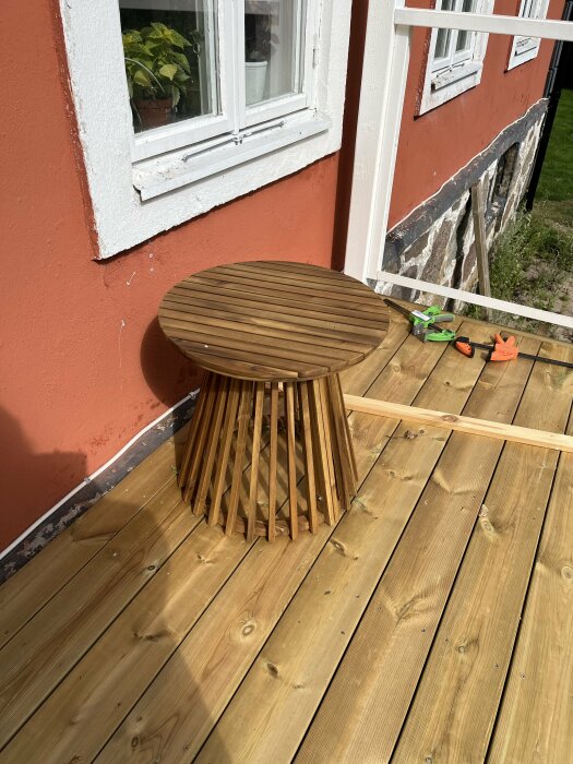 Litet sidobord i trä monterat på en veranda med trallbrädor, intill ett fönster på ett hus med tegelröd fasad.