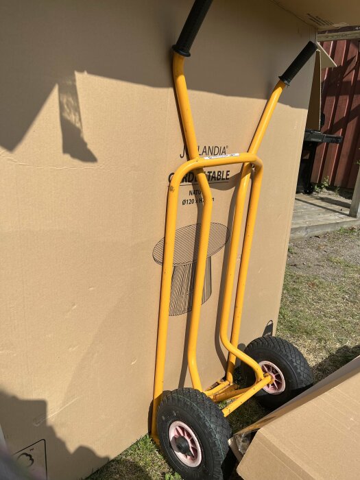 Handtruck lutad mot stor kartong med texten "Jutlandia Garden Table" på gräsmatta utomhus.