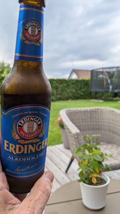 En person håller en flaska alkoholfri Erdinger Weissbräu på en uteplats med trädgårdsmöbler och häck i bakgrunden.