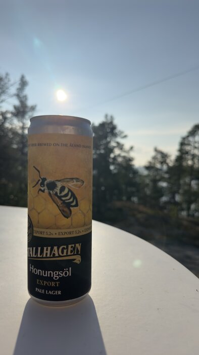 En burk med Tallhagen Honungsöl Export Pale Lager står på ett bord utomhus med natur i bakgrunden och solen på väg ner.