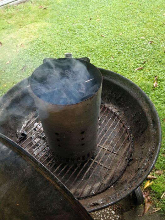 En kolgrill med ett rökkärl fullt med brinnande kol står på en grön gräsmatta. Grillgallret är sotigt och kolen ryker.