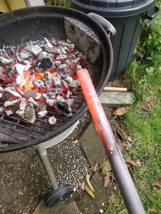 En glödande metallstång hålls över en grill fyllt med heta kolbitar, med en svart soptunna och gräs i bakgrunden.