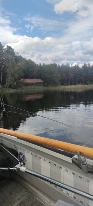 En stuga i skogen vid en sjö, sedd från en båt med en fiskespö i förgrunden och en molnig himmel i bakgrunden.