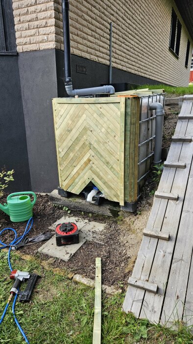 Inbyggd IBC-tank med träpanel bredvid ett hus, omgiven av verktyg och en grön vattenkanna på marken.