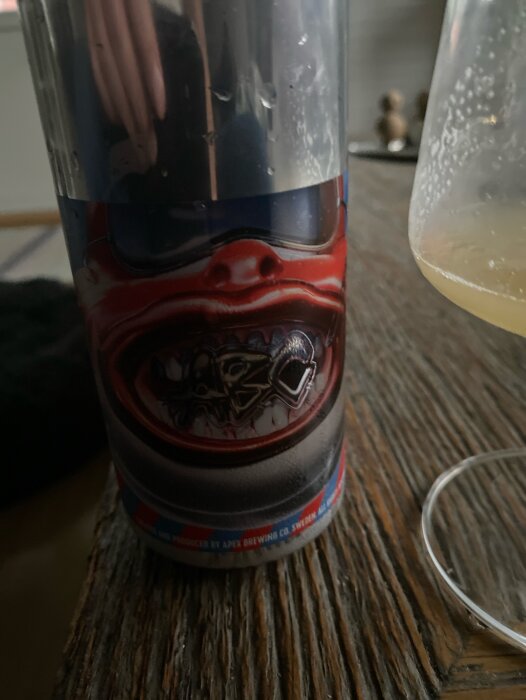 En burk Apex Helios IPA öl stående på ett träbord bredvid ett glas med öl.