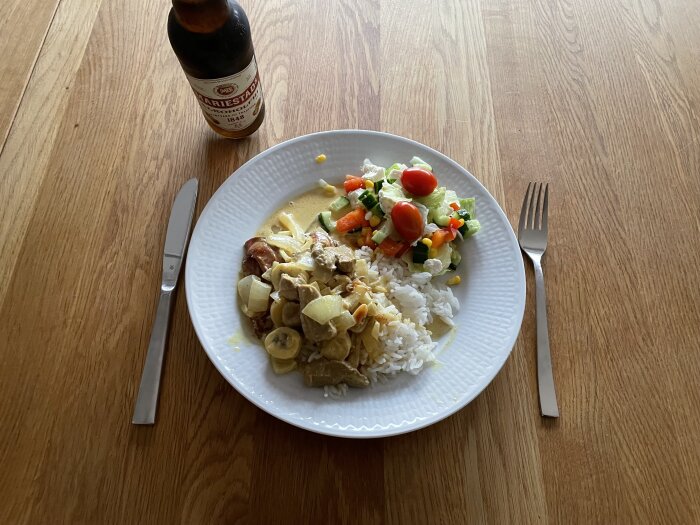 Porslinstallrik av fläskfilé Africana med ris och sallad, omgiven av en gaffel, en kniv och en Mariestads ölflaska på ett träbord.