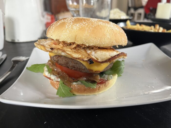 Hamburgare med stekt ägg, rostad och stekt lök, ost, tomat, sallad, gurkmayonnaise, sirrachadressing och nötkött serverad på ett vitt hamburgerbröd.