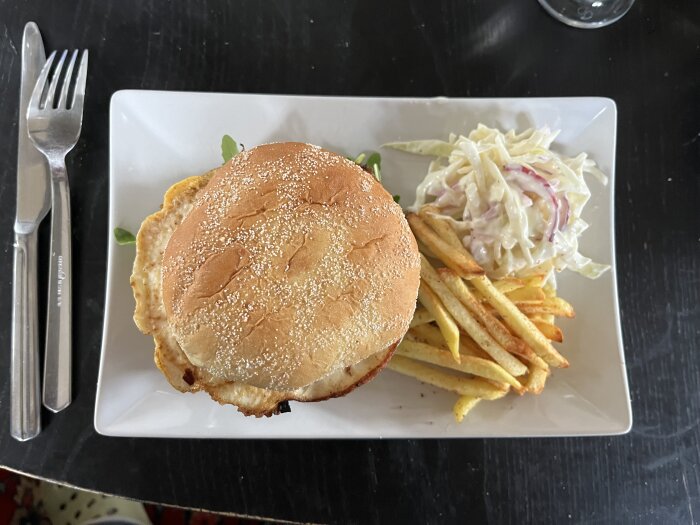 Hamburgare med stekt ägg serverad på en tallrik med pommes frites och coleslaw, tillsammans med bestick på en mörk bordsyta.