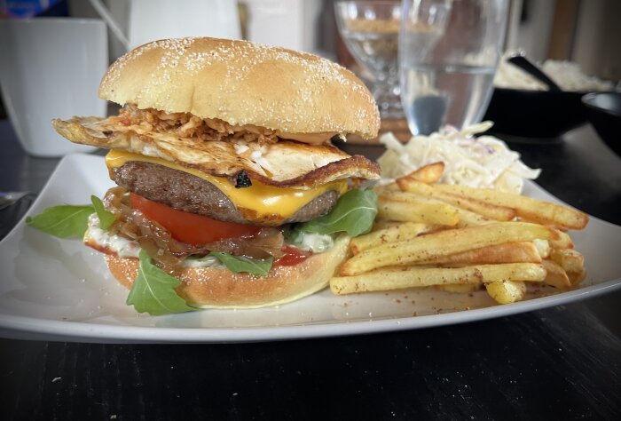 Hamburgare med stekt ägg, ost, stekt lök, grönsaker och dressing på en tallrik, serverad med pommes frites och coleslaw i bakgrunden.