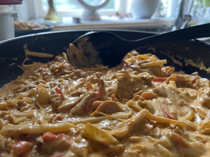 En wokpanna fylld med thai-fusionrätt innehållande kyckling, kokosmjölk, currypasta, bambuskott, paprika, vitkål, vitlök och lök, med en svart slev i rätten.