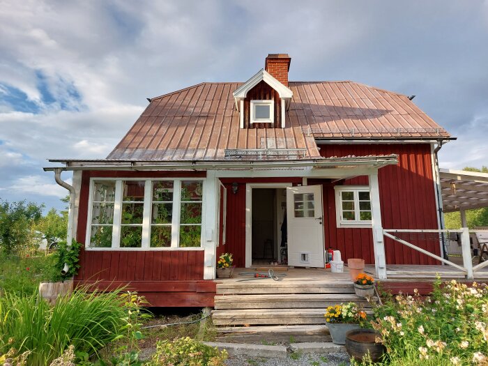 Rött hus med plåttak och vit dörr, sett från framsidan. Trädgårdsblommor i förgrunden, stege och byttor. Skorsten på toppen av taket, omgiven av grönska.