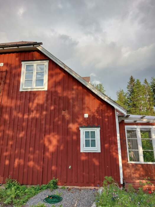 Rött hus med vitmålade fönster och ett plåttak. Trädens skuggor syns på fasaden. Vattenkran och en hoprullad trädgårdsslang på marken.