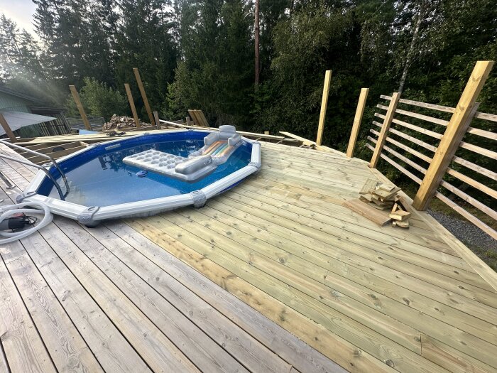 Trädäck i byggprocess med flexibel pool och uppblåsbar madrass, omgärdat av träribbor och omgivande skog. Verktyg och material ligger på trädäcket.